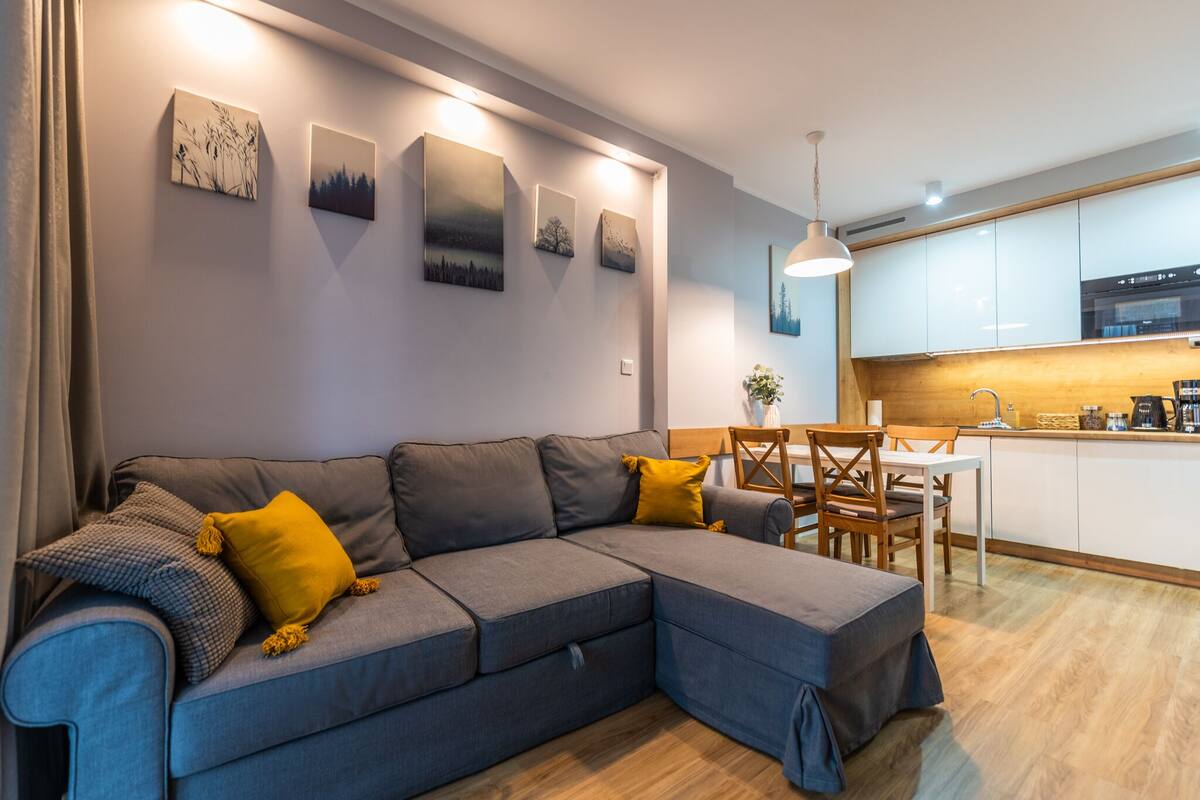 Un salon moderne avec un canapé d’angle gris, une table à manger et un coin cuisine doté de meubles blancs et d’un plan de travail en bois.