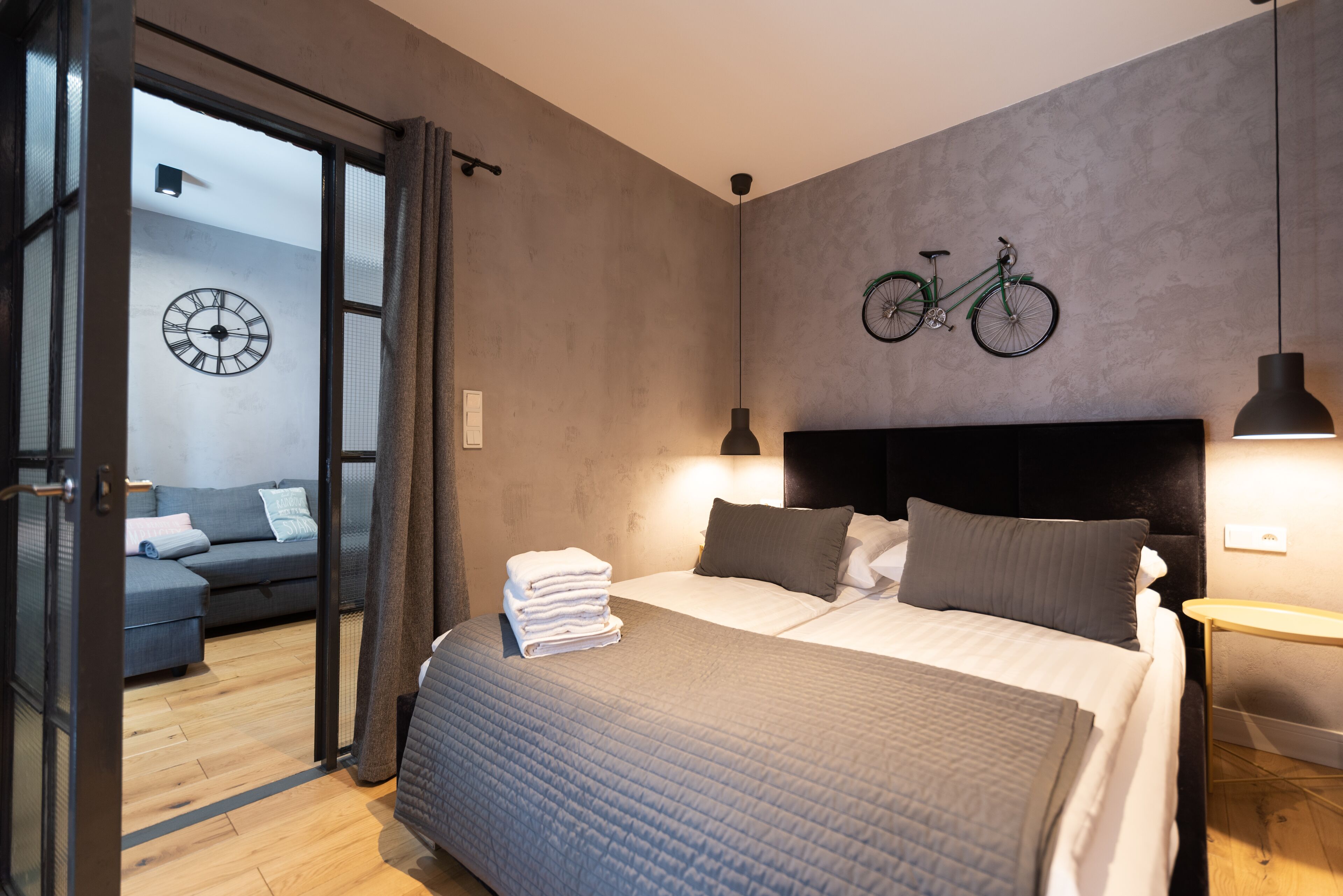 A modern bedroom with a bed, bedside tables, a bicycle wall art, a clock, and a view into another room with a sofa.
