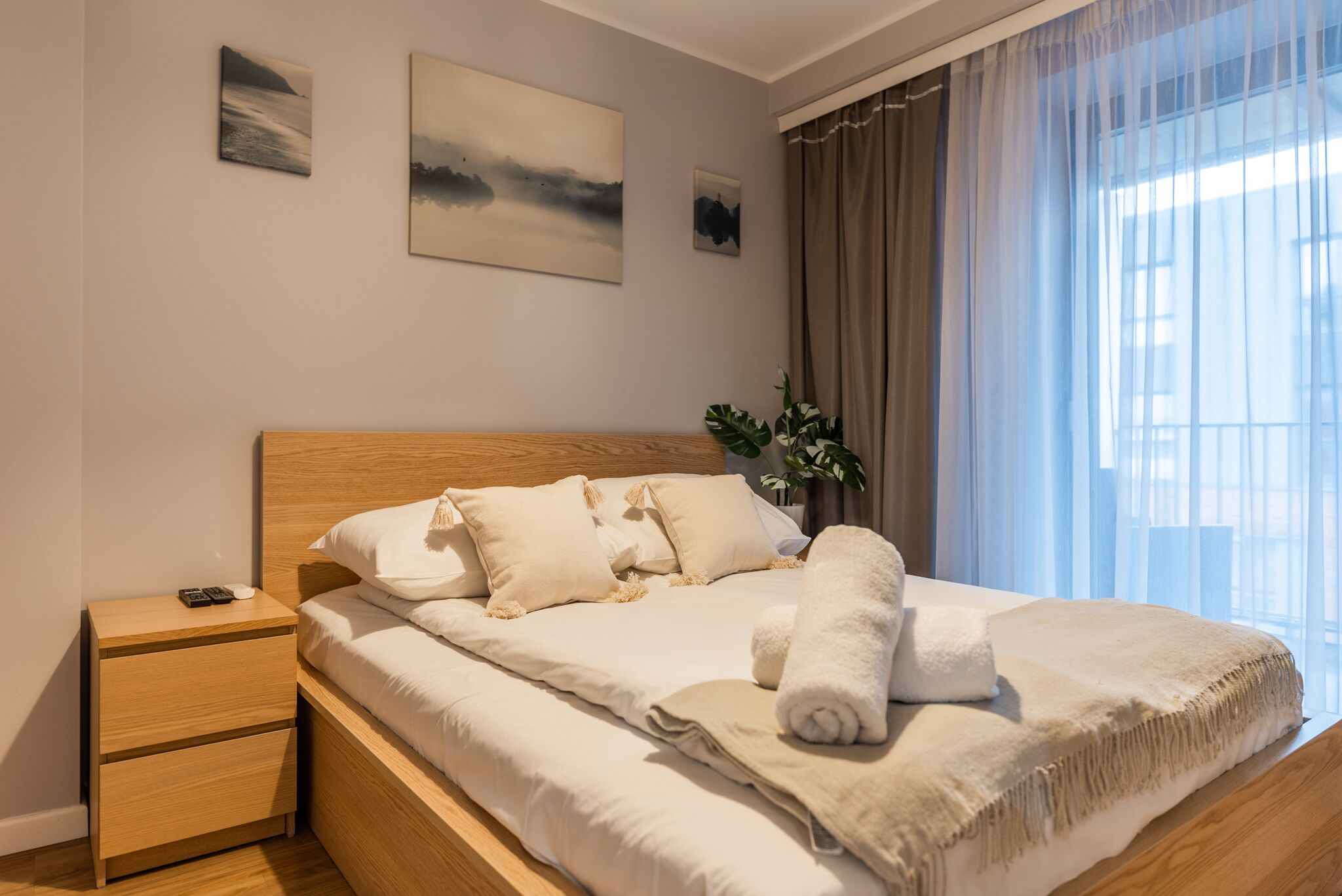 A neatly made bed with a wooden headboard, a bedside table with a lamp, and a window with sheer curtains.