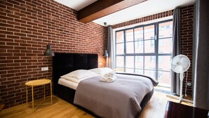 A modern hotel room with a brick wall, a large window, a bed with a black headboard, a bedside table, and a standing fan. - City Lights Kazimierz (Kraków)