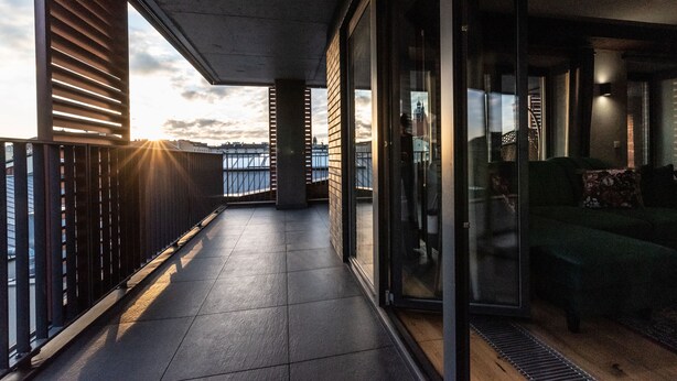 A balcony with a view of the sunset, a glass door leading to a living area with a sofa and a coffee table. - City Lights Kazimierz (Kraków)