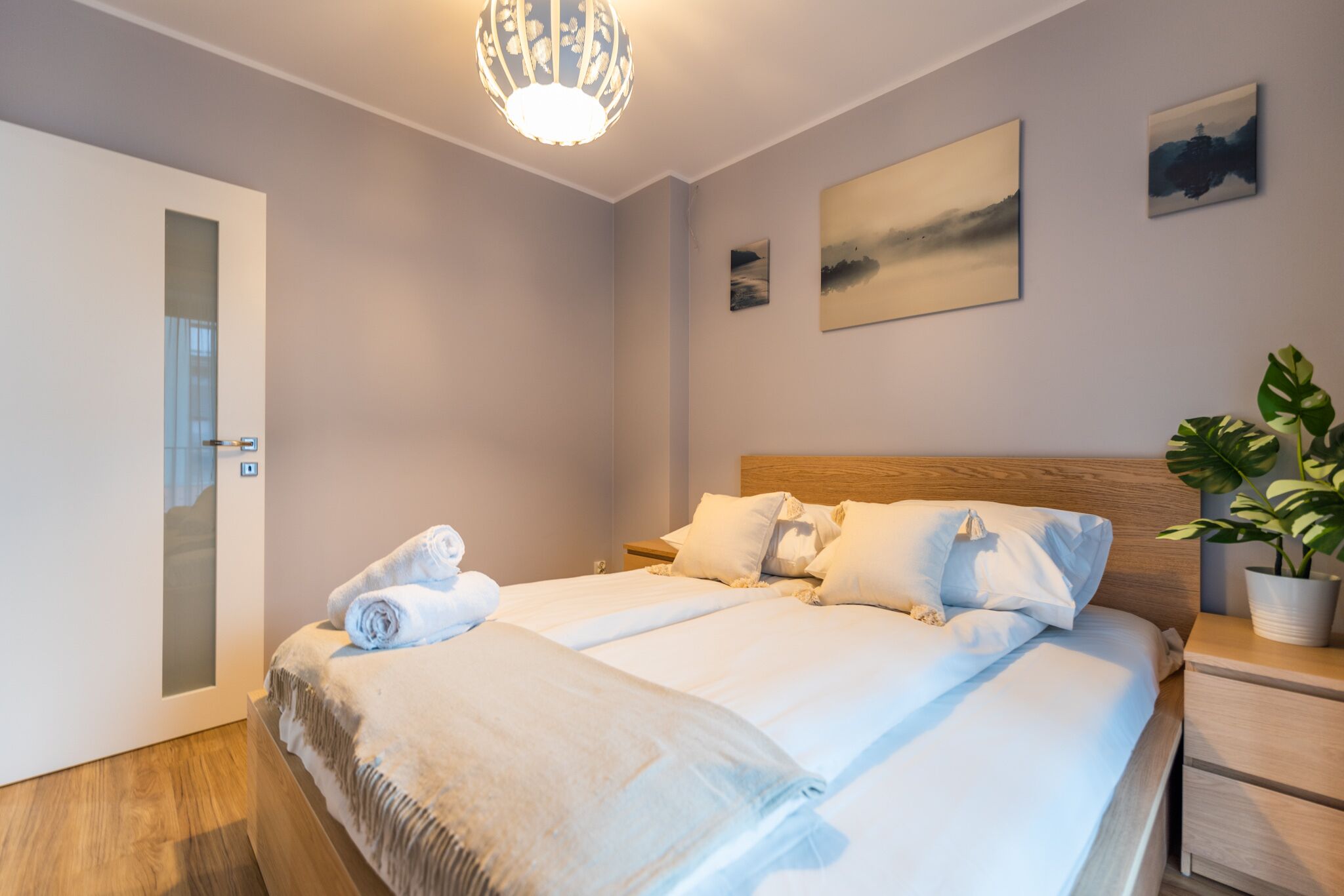 A neatly made double bed with white linens, a wooden headboard, and a bedside table with a potted plant.