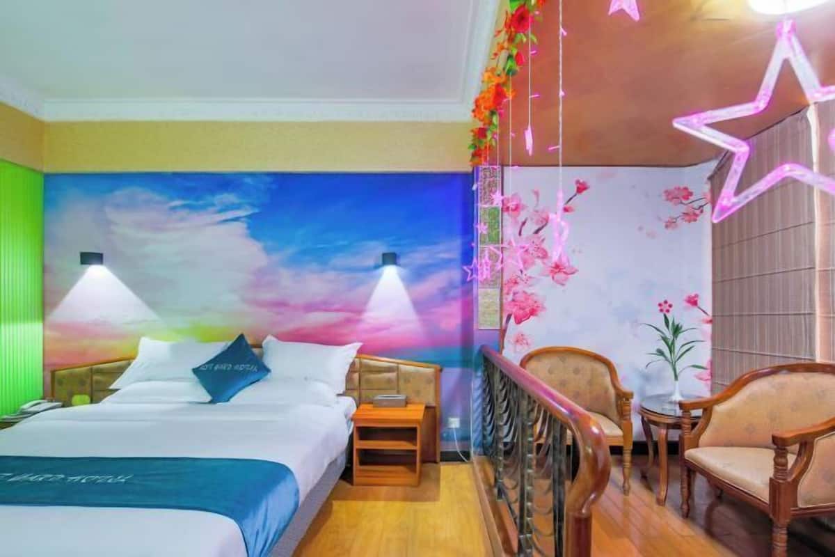 A hotel room with a bed, bedside tables, a nightstand, a chair, a small table, and a decorative wall with floral patterns.
