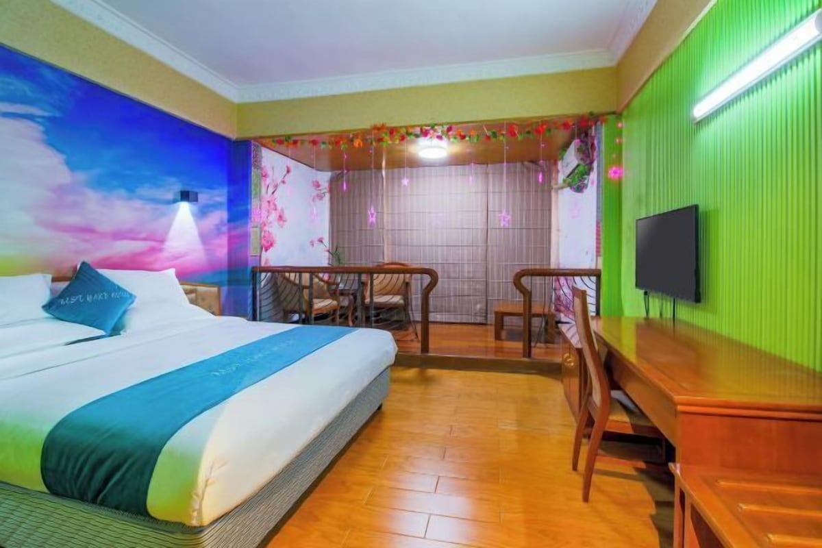 A hotel room with a bed, a desk, a chair, a TV, and a mural on the wall.