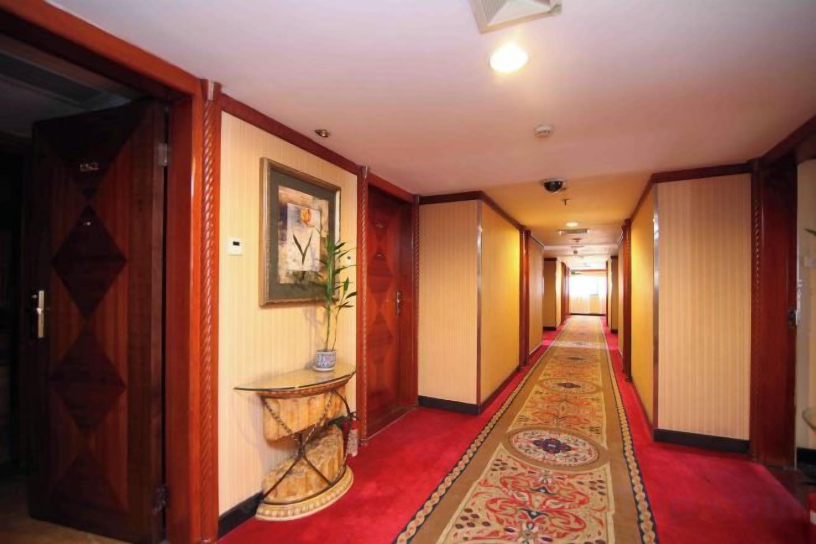 A hotel corridor with a red carpet, wooden doors, a framed picture, and a plant on a console table.