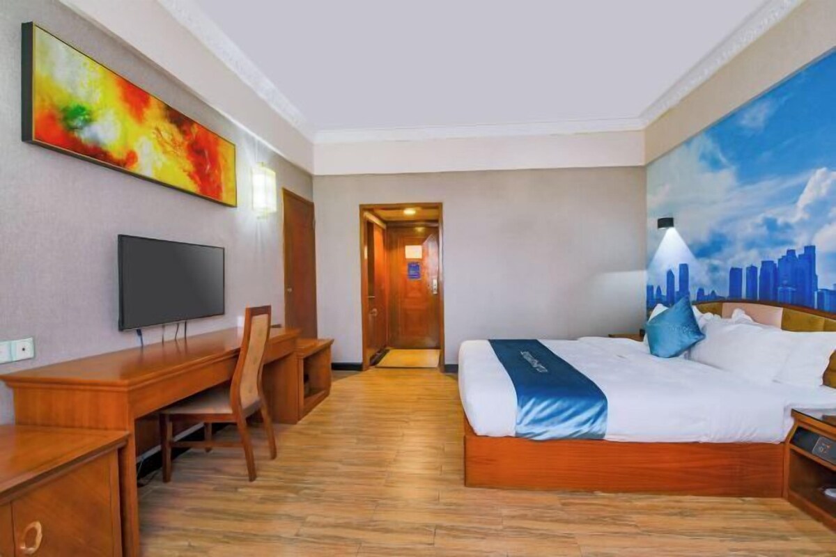 A hotel room with a bed, a desk, a chair, a television, and a painting on the wall.