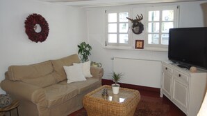 A living room with a beige sofa, a wicker coffee table, a television on a white cabinet, and a window with a wreath and a deer head decoration. - The small Villa OLEFant in the historic center of Schleiden Olef (Schleiden)