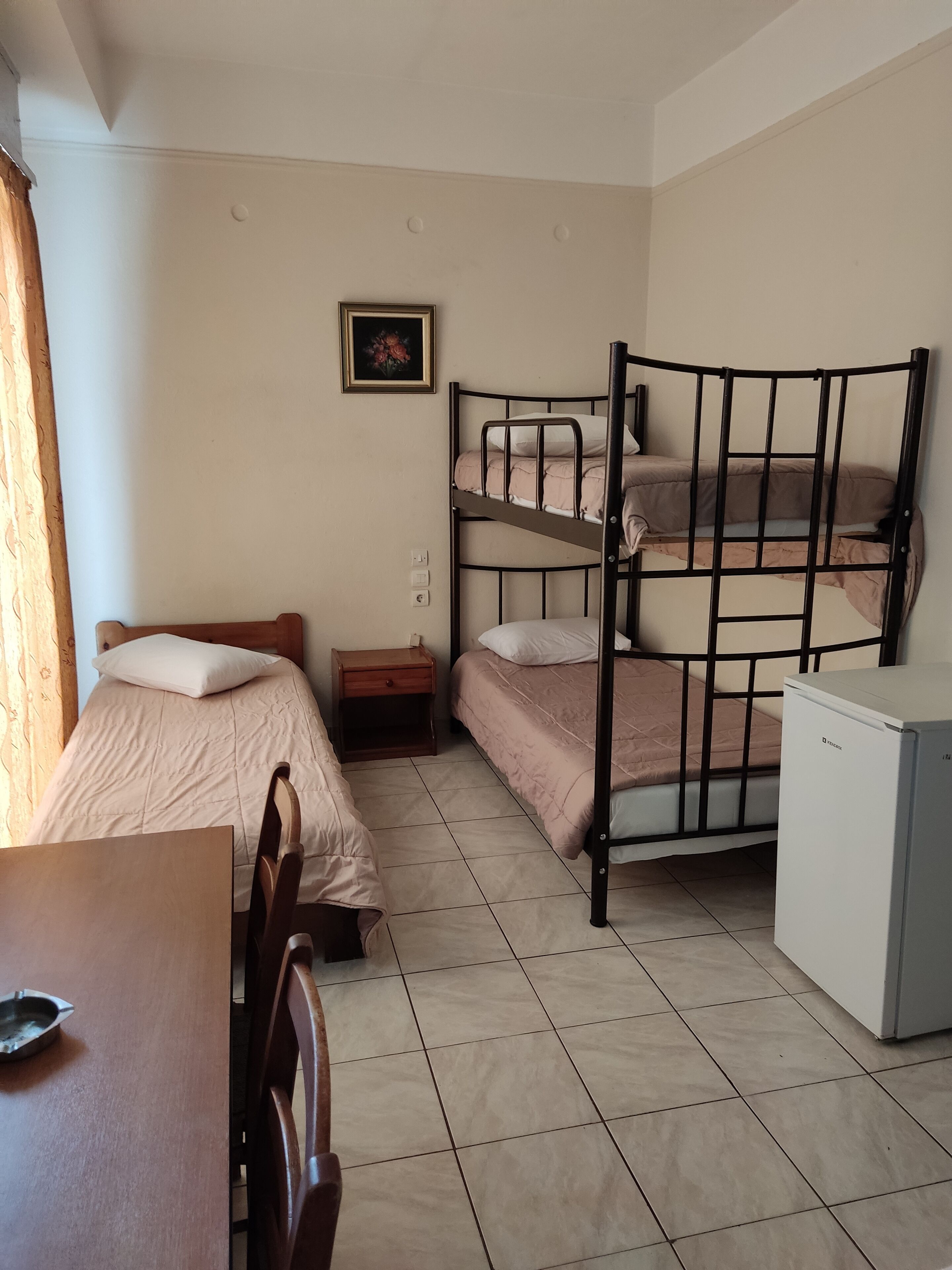 Une chambre de dortoir avec des lits superposés, un bureau, une chaise, une petite table, une table de chevet, un réfrigérateur et un tableau encadré au mur.