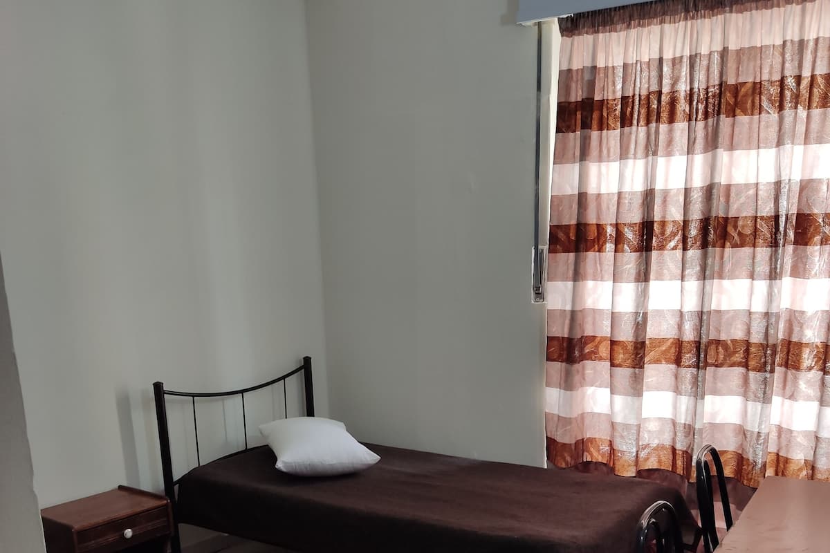 A small, single-room accommodation with a bed, a bedside table, a chair, and a desk.