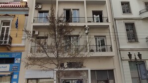 A multi-story building with balconies, a storefront on the ground floor, and a tree in front.
