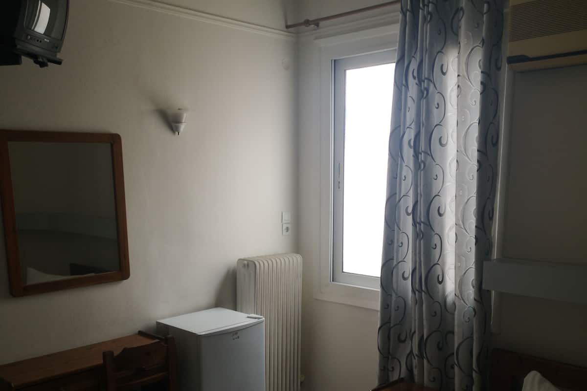 A small, simple room with a bed, a desk, a radiator, and a window with curtains.