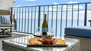 A balcony with a glass table displaying a bottle of wine, two glasses, and a platter of fruits and crackers.
