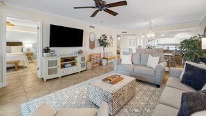 A spacious living room with a ceiling fan, a large flat-screen TV, a grey sofa, and a coffee table.