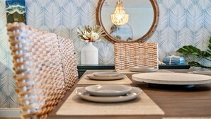 A dining area with a wooden table set for four, a woven chair, a mirror, and a hanging light fixture.