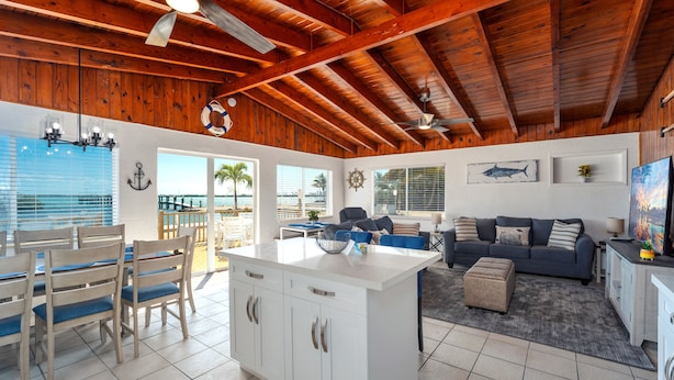 Un espacio habitable amplio con techo de madera, una isla grande con muebles blancos, un sofá gris y un comedor con sillas y mesa de madera.