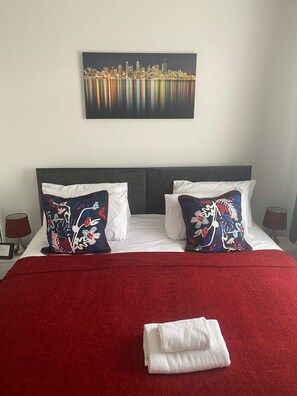 A hotel room with a large bed, two decorative pillows, a red blanket, and a cityscape painting on the wall. - Remarkable 3-bed House in Ipswich (Ipswich)