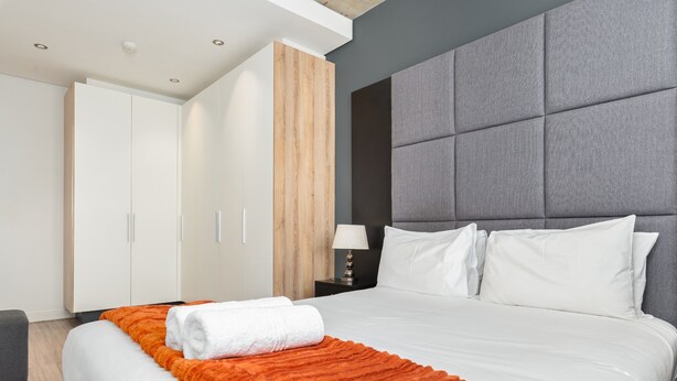 Una habitación de hotel moderna con una cama grande, un cabecero acolchado gris, ropa de cama blanca y una pared decorativa de madera.