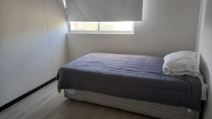 A small, single-bed room with a plain white wall, a window with a white blind, and a mattress with a quilted cover.