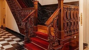 Una imponente escalera con detalles de madera ornamentales y peldaños alfombrados de rojo, que conduce a un rellano con una puerta blanca que indica "DUKE".