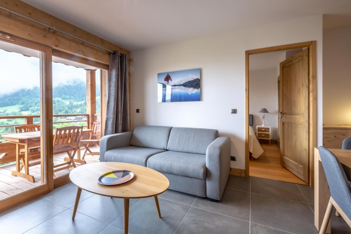 A modern living room with a grey sofa, a round wooden table, and a balcony with wooden furniture.
