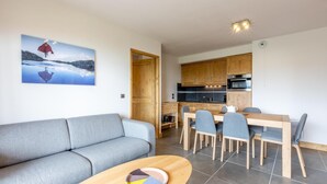 Un salon moderne avec un canapé gris, une table à manger en bois avec quatre chaises, et un coin cuisine doté de meubles en bois et d’un plan de travail noir.