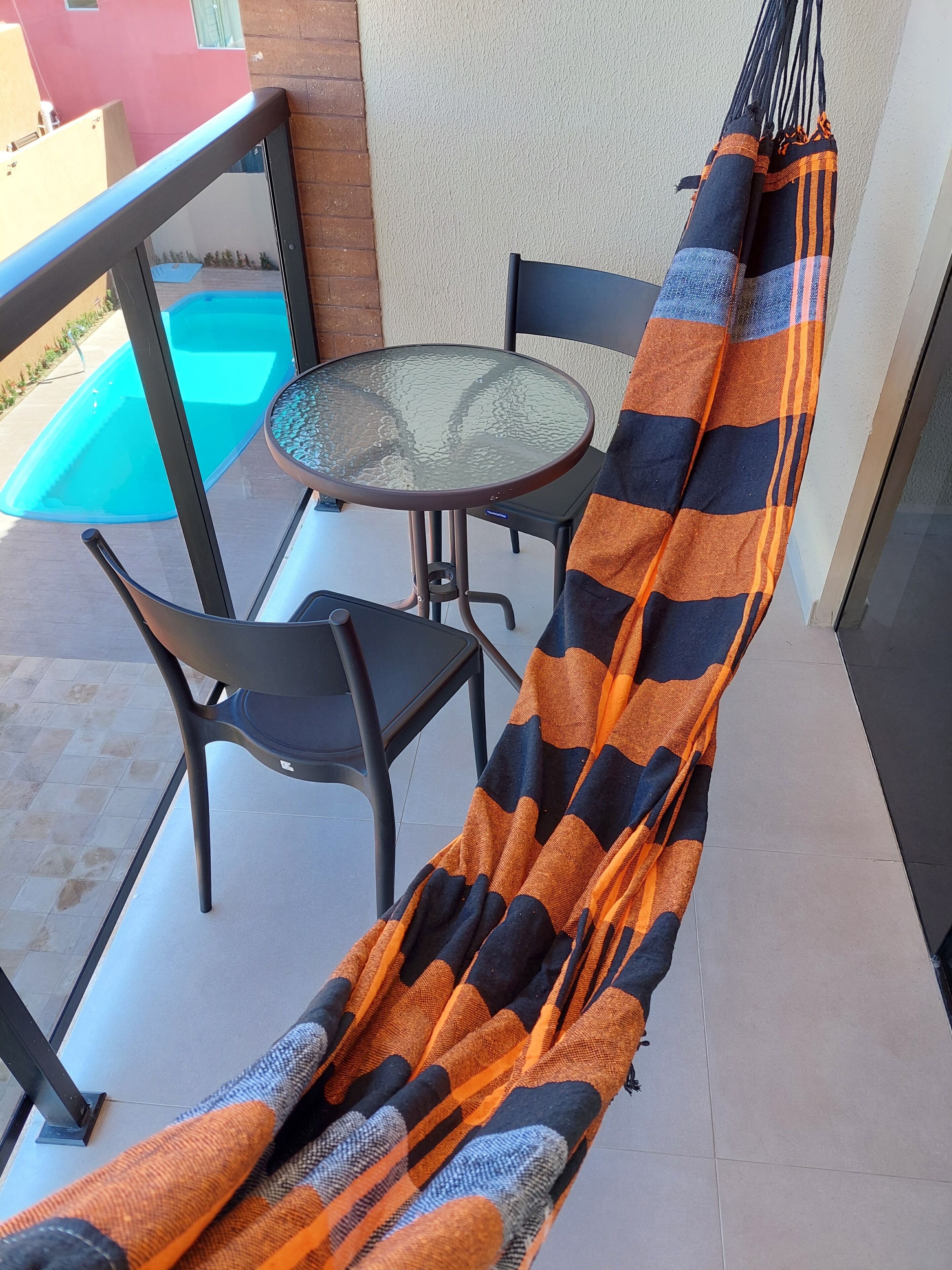 Un'amaca su un balcone con un tavolo in vetro e sedie, una piscina sullo sfondo e un pavimento piastrellato.