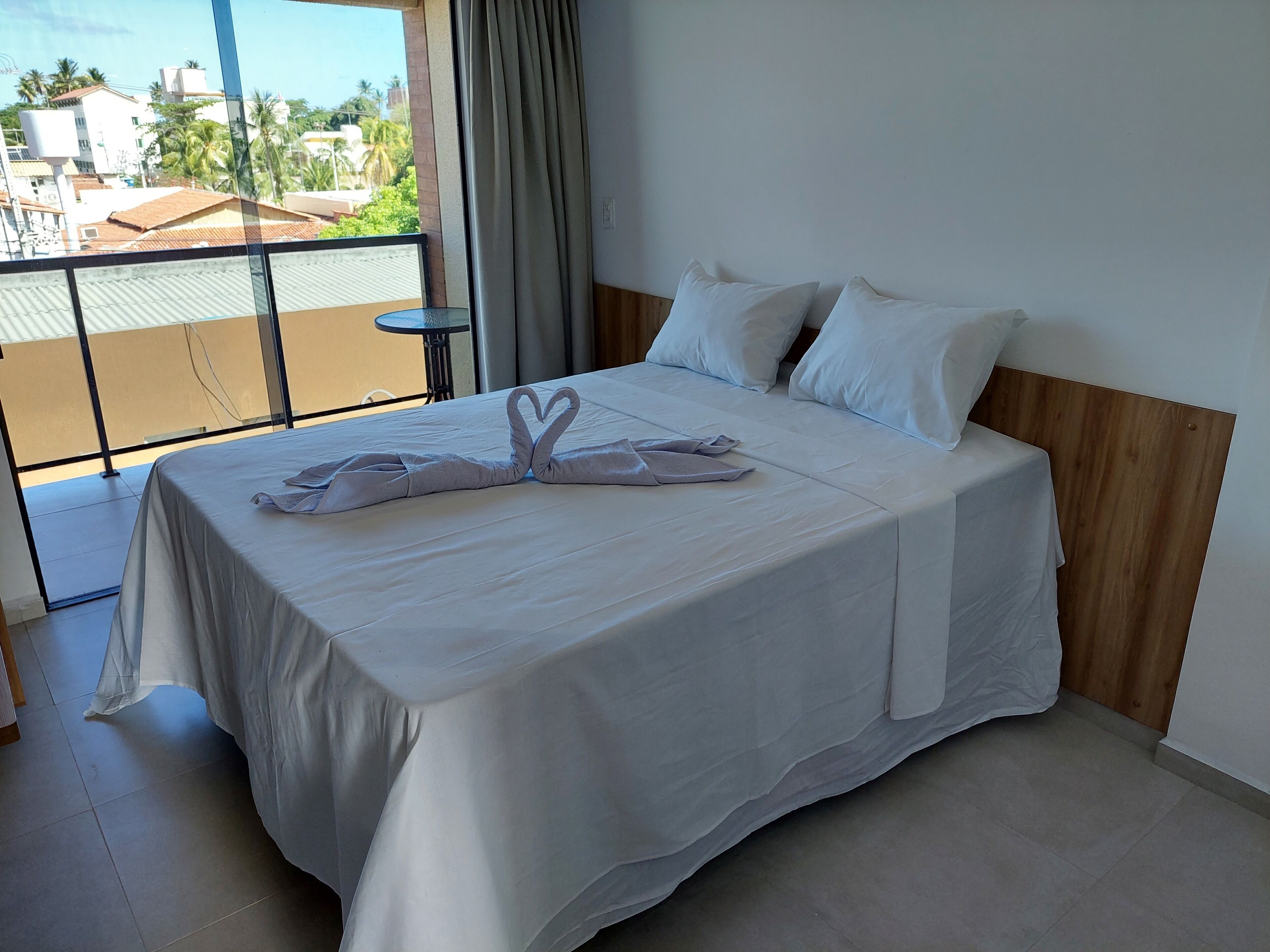 A hotel room with a large bed, white linens, a wooden headboard, and a view of buildings and palm trees through a balcony.