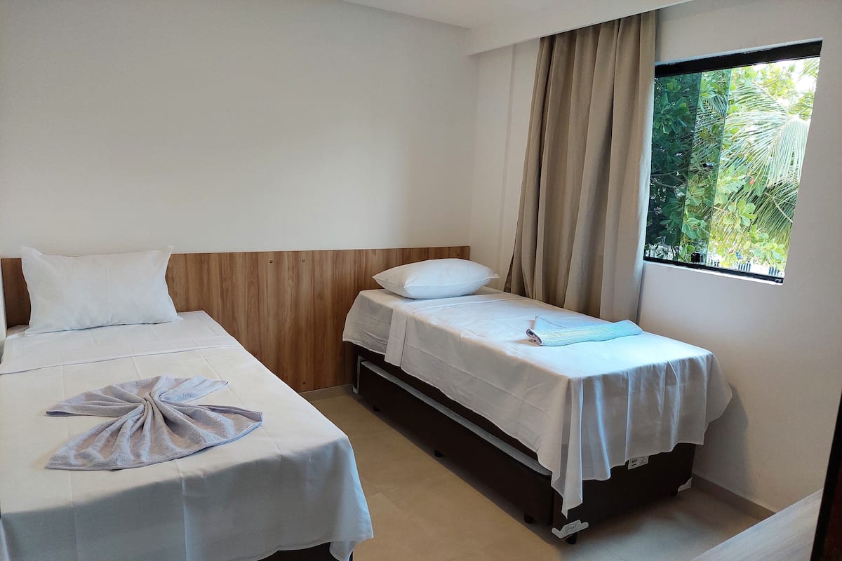 A hotel room with two beds, a window with curtains, and a view of greenery outside.