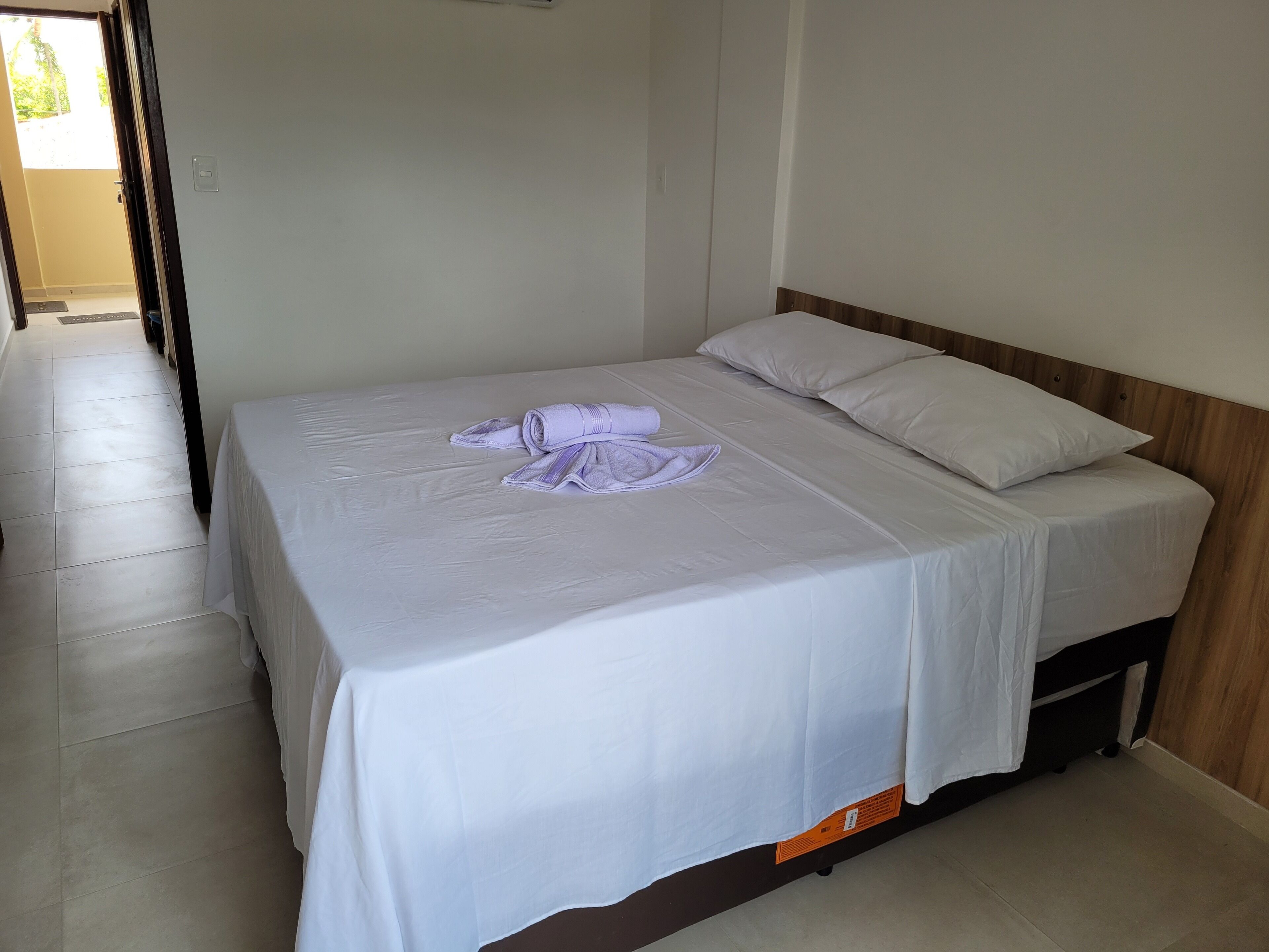A neatly made bed with white linens and a rolled towel on top, a wooden headboard, and a plain white wall.