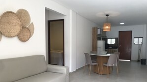 Una sala de estar moderna con un sofá, una mesa de comedor y sillas. Se aprecia una cocina con armarios y un refrigerador al fondo.