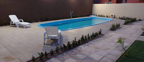 A swimming pool with two lounge chairs and a small table, surrounded by a tiled patio and a low hedge.