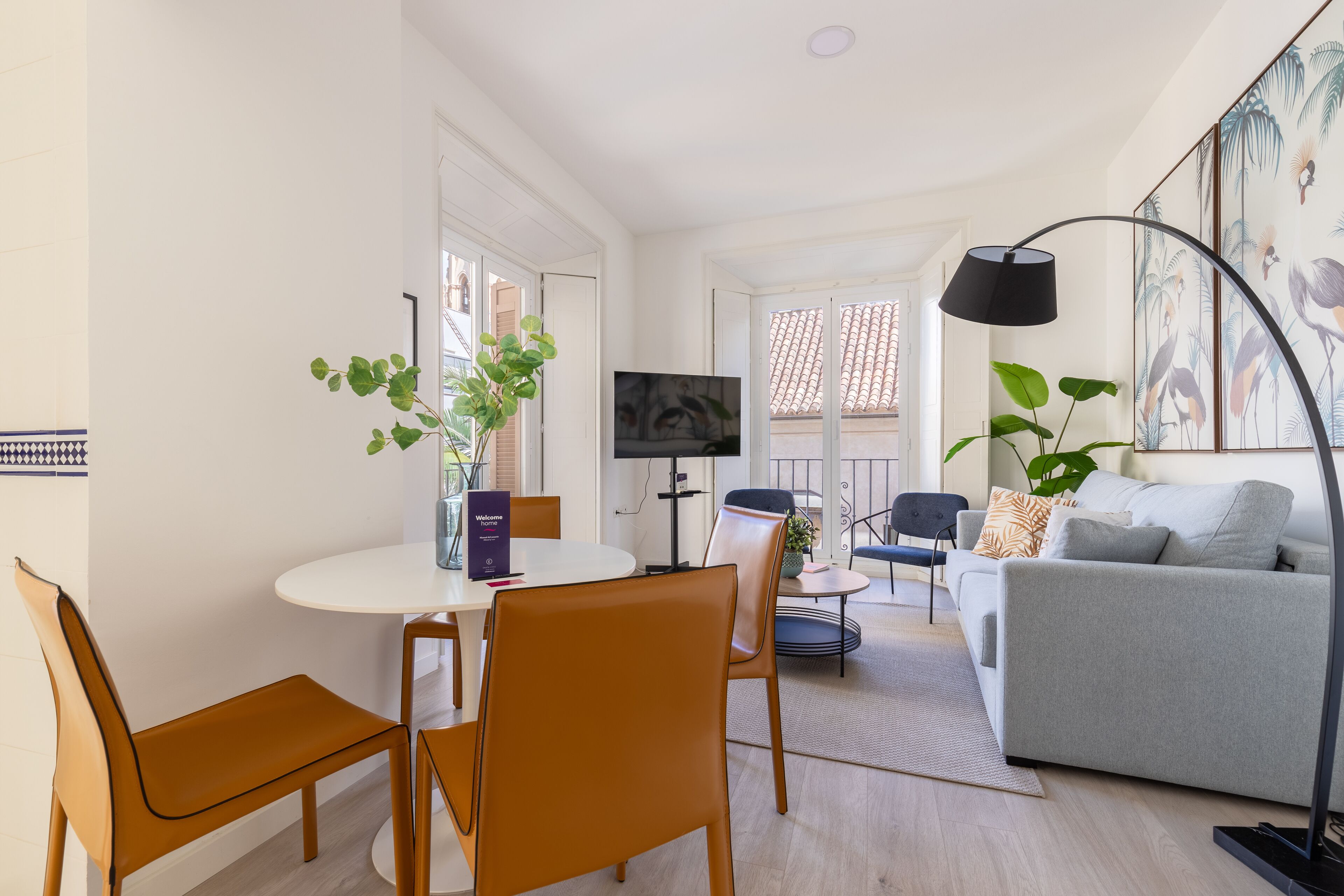 A modern living room with a white sofa, a grey couch, a round dining table with chairs, a TV, and a large plant.