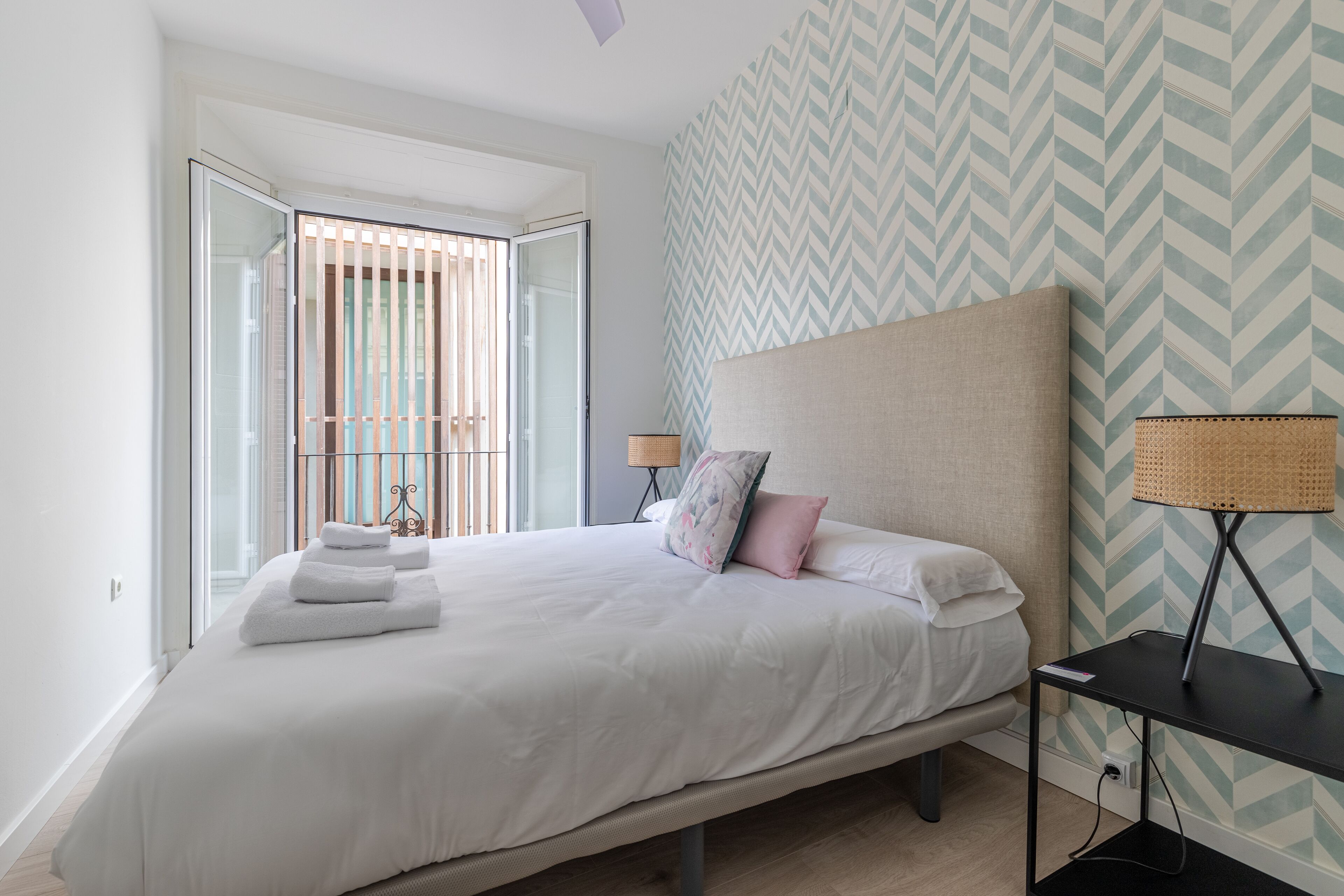 Een netjes opgemaakt bed met witte lakens en een paar kussens, een nachtkastje met een lamp, en een behang met een patroon.