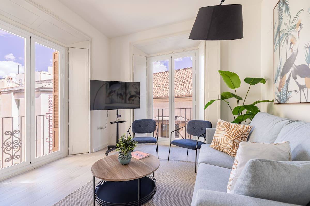 Ein modernes Wohnzimmer mit einem grauen Sofa, einem runden Couchtisch und einem Fernseher auf einem Schrank. Ein Balkon bietet Blick auf Gebäude, auf dem Couchtisch steht eine Pflanze.