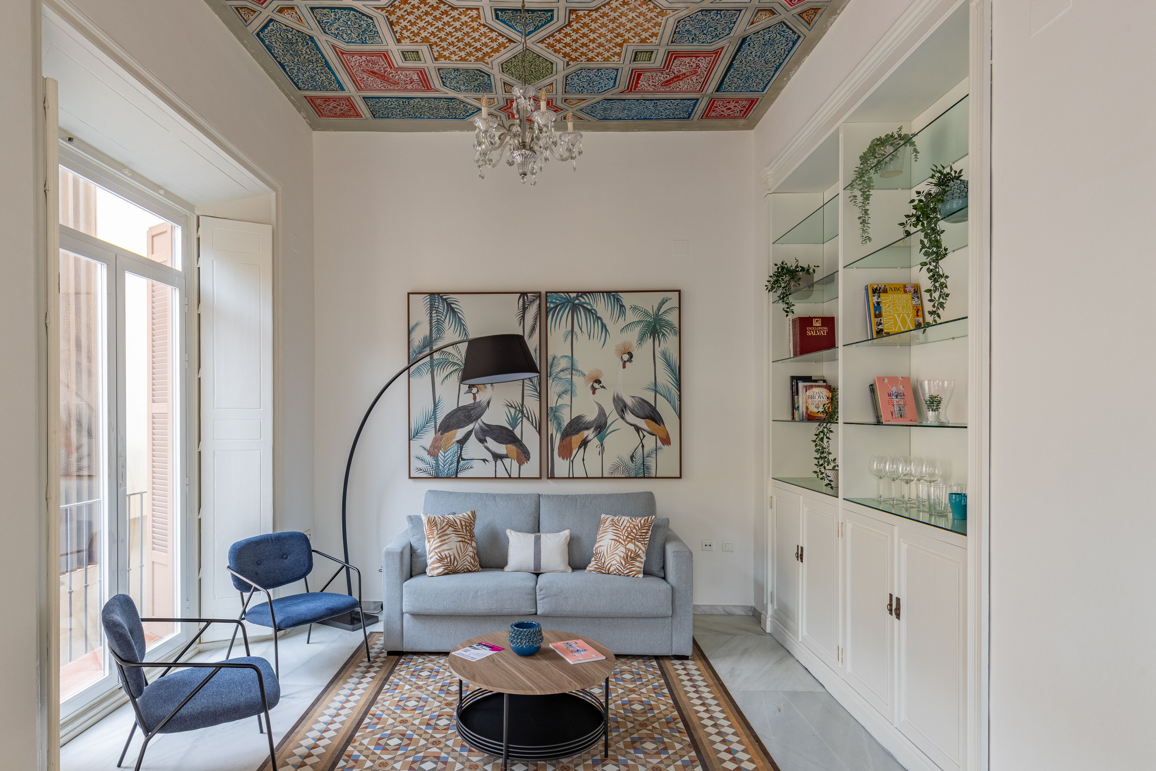 A living room with a light grey sofa, two blue armchairs, a patterned rug, a round coffee table, and a bookshelf with glass doors and decorative items.