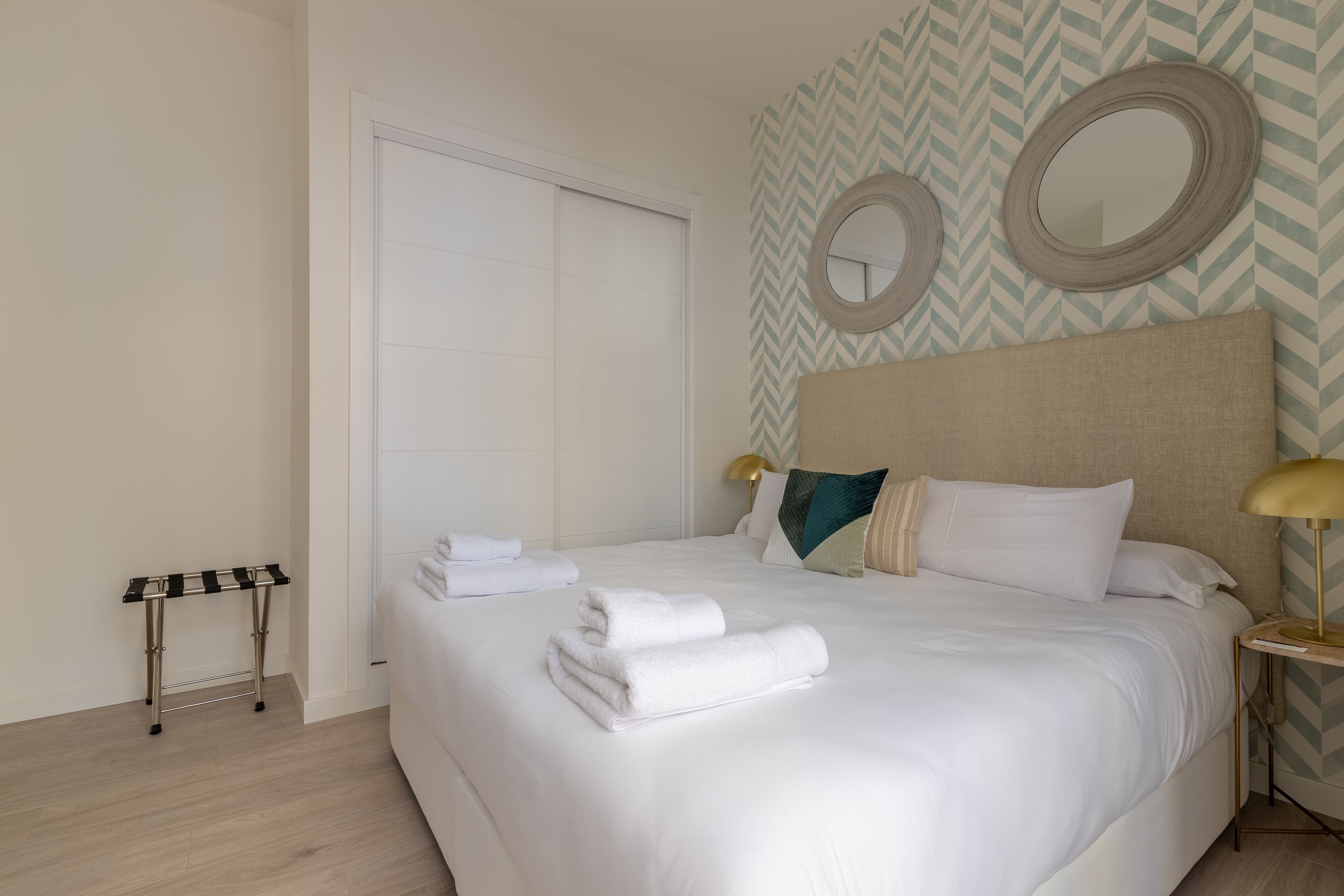 Un letto rifatto con lenzuola bianche e asciugamani piegati, un comodino con una lampada, e una parete con specchi decorativi e carta da parati a motivi a zig-zag.