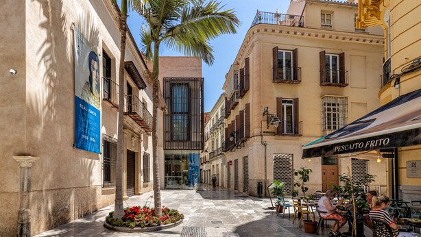 Une rue étroite, bordée de bâtiments des deux côtés, avec des tables en terrasse devant un restaurant et un palmier.
