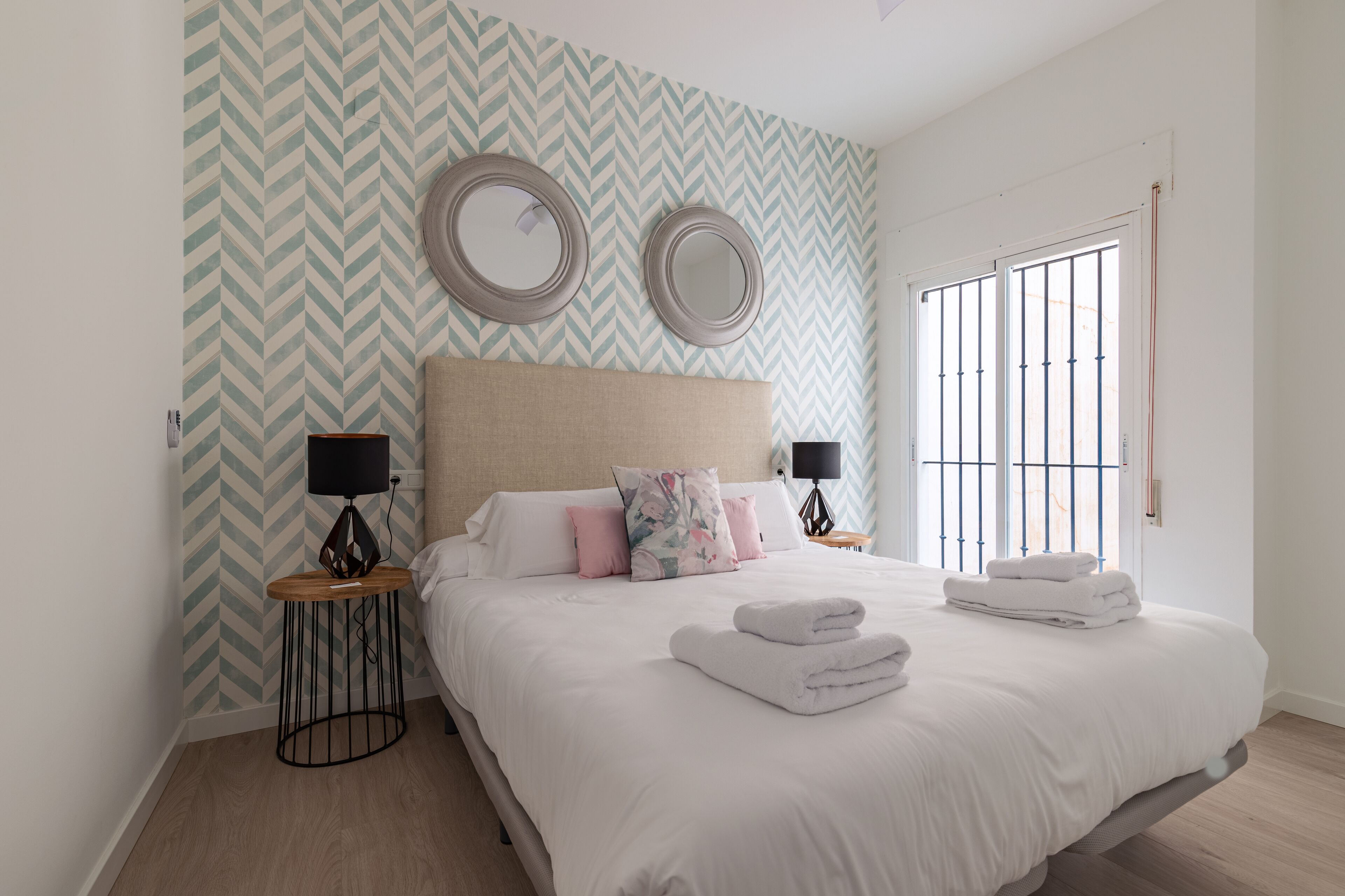 A neatly arranged bedroom with a large bed, two bedside lamps, a mirror, and a window with bars.