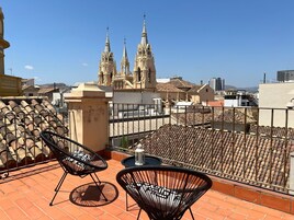 Una terraza en la azotea con dos sillas de alambre negras y una mesa pequeña, con vistas a una iglesia histórica de tres torres.