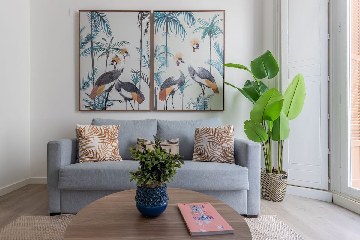 Ein Wohnzimmer mit grauem Sofa, gemusterten Kissen, einer blauen Vase mit Pflanze und einem gerahmten Bild von Vögeln an der Wand.