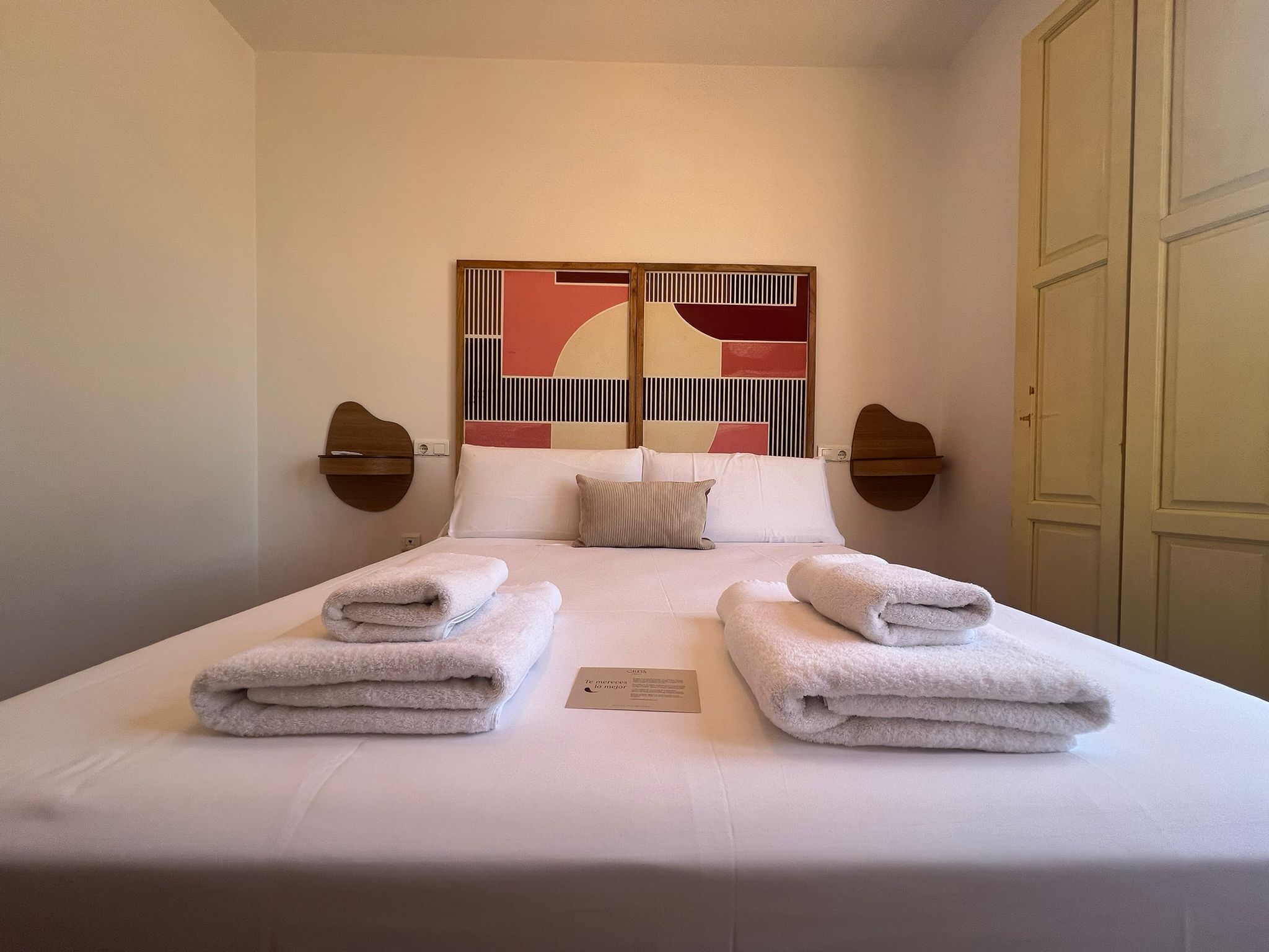 A hotel room with a bed, two bedside tables, a wall art piece, and a door.