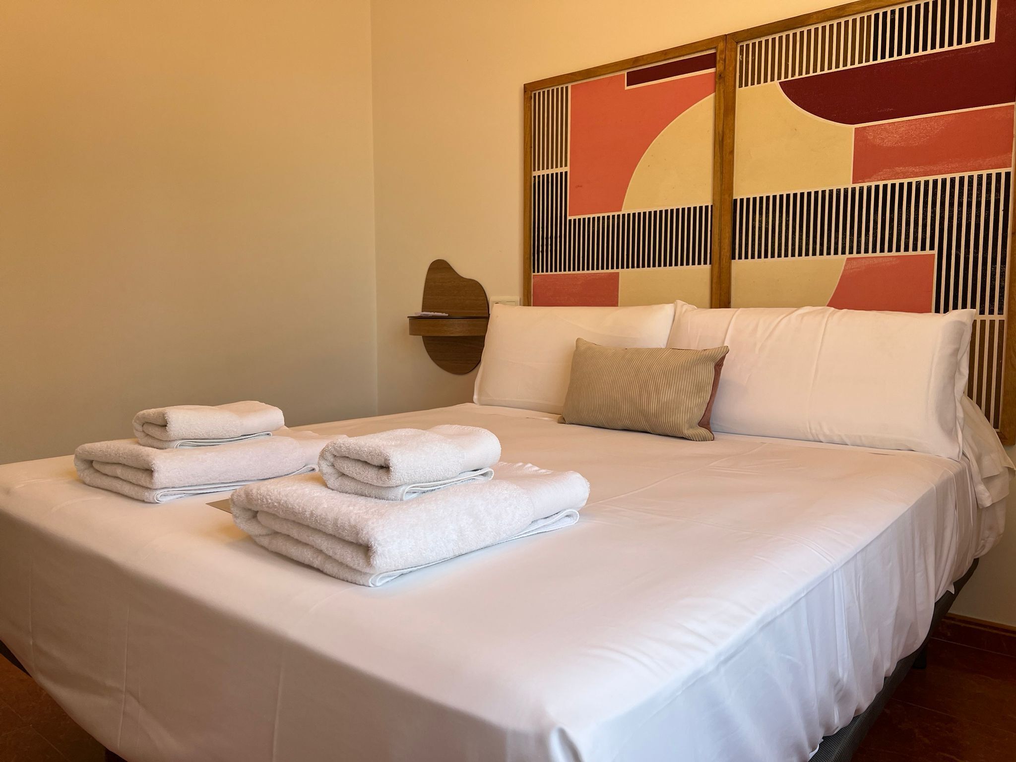 A neatly made bed with white linens and folded towels, a headboard with a geometric pattern, and a wall-mounted light fixture.