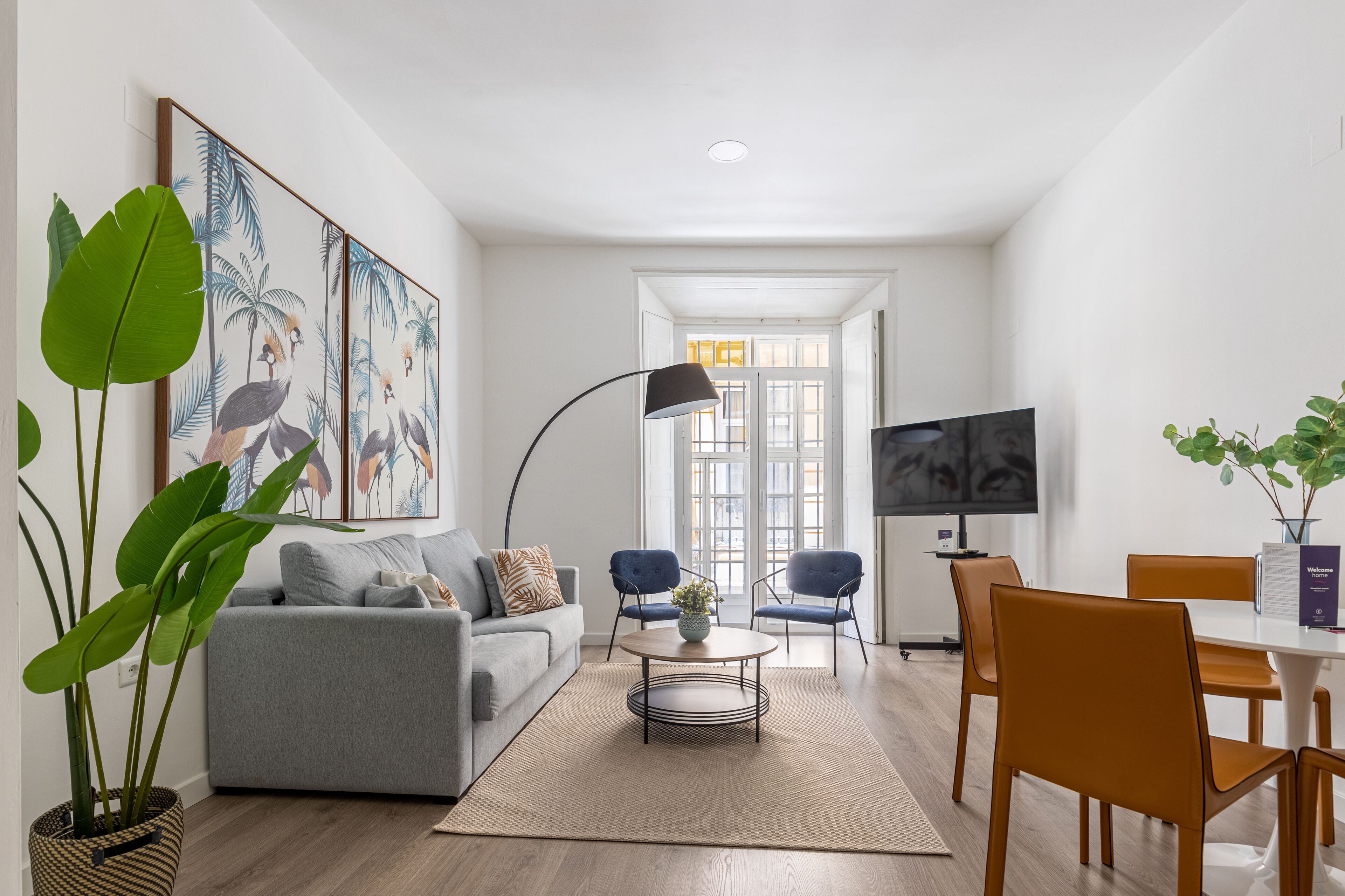 Una sala de estar moderna con un sofá gris, una mesita de centro y un televisor. Hay dos sillones y una mesa de comedor con sillas naranjas. Una planta grande está en la esquina, y hay cuadros en la pared.