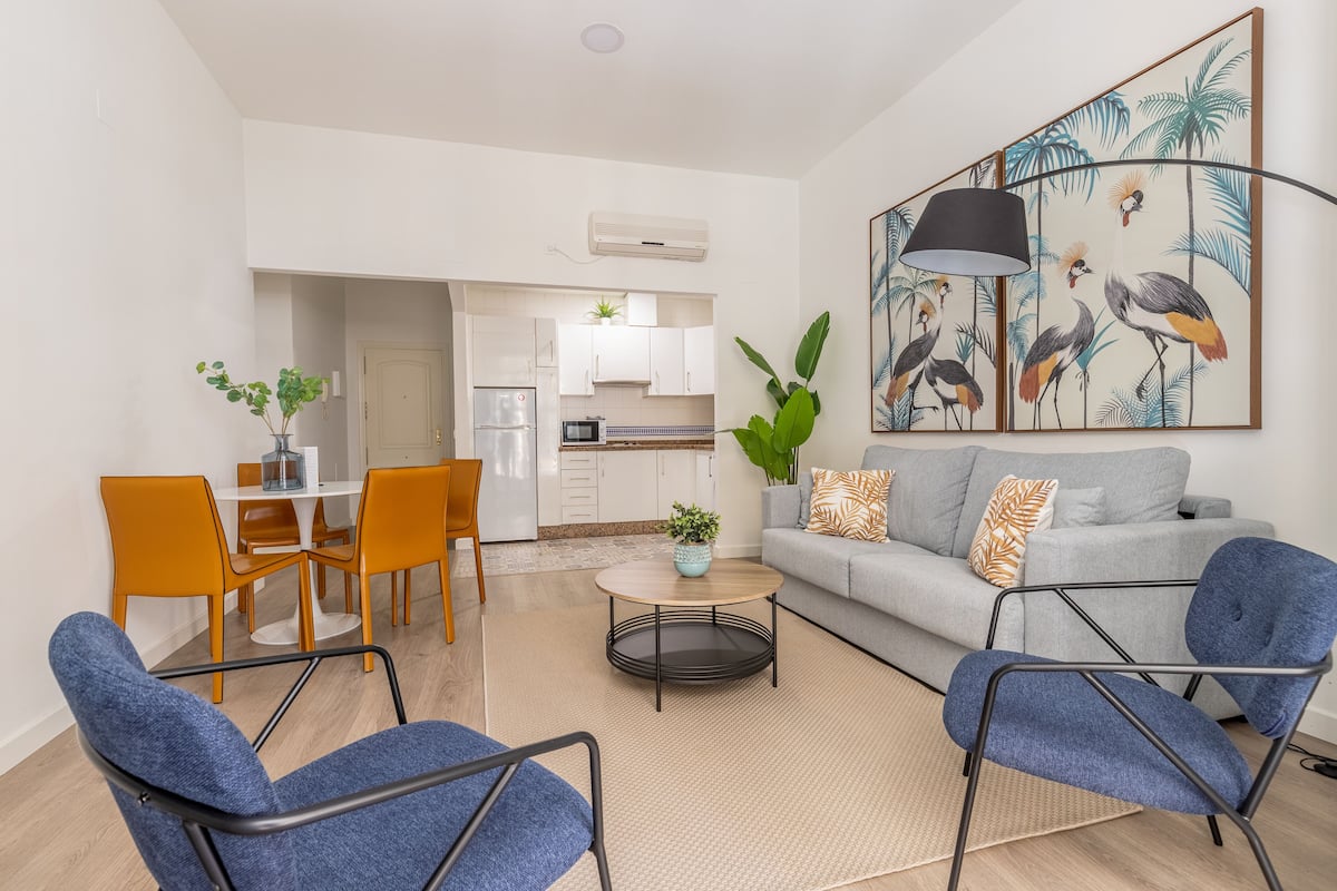 Ein modernes Wohnzimmer mit einer grauen Couch, blauen Sesseln und einem Couchtisch. Es gibt eine Essecke mit orangefarbenen Stühlen und einem weißen Tisch. Im Hintergrund ist eine Küche mit weißen Schränken und einer Mikrowelle zu sehen.