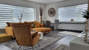 A modern living room with a sofa, armchair, and ottoman, a white coffee table, and a large window with blinds. - Lounge 2 - Top apartment approx. 72sqm, air conditioning, parking at the house, all in (Koblenz)