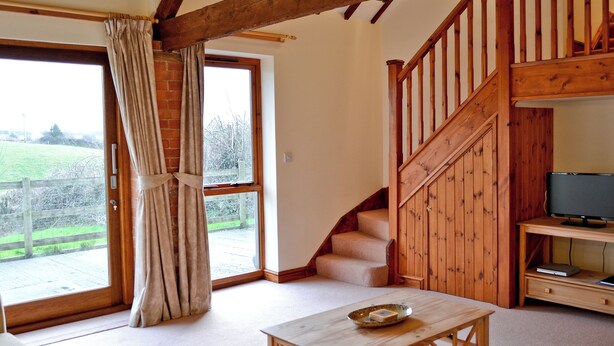 A spacious living room with a wooden staircase, a television on a wooden stand, and a view of a green landscape through large windows.