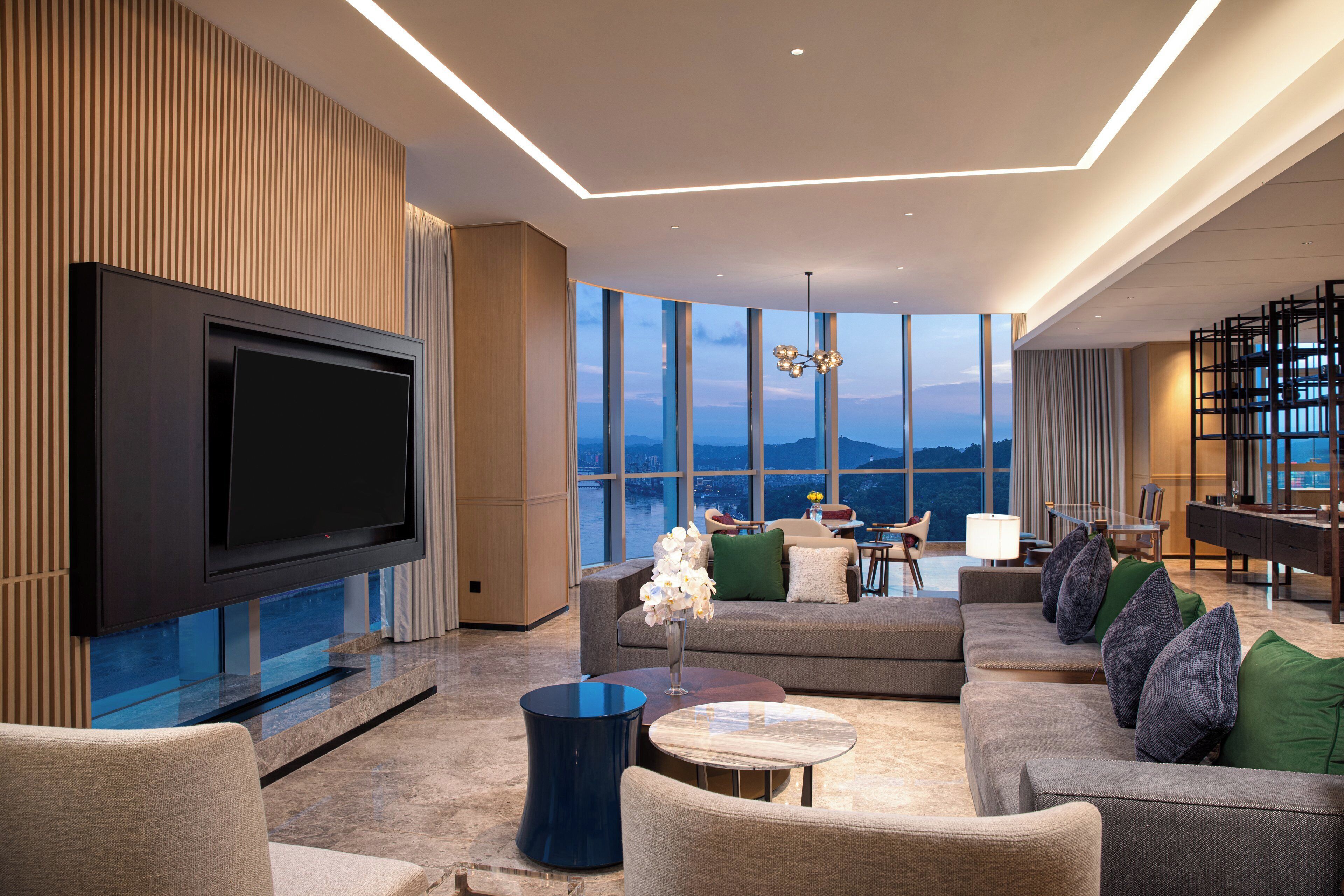 A modern living room with a flat-screen TV, comfortable seating, a coffee table, and a view of the cityscape through large windows.