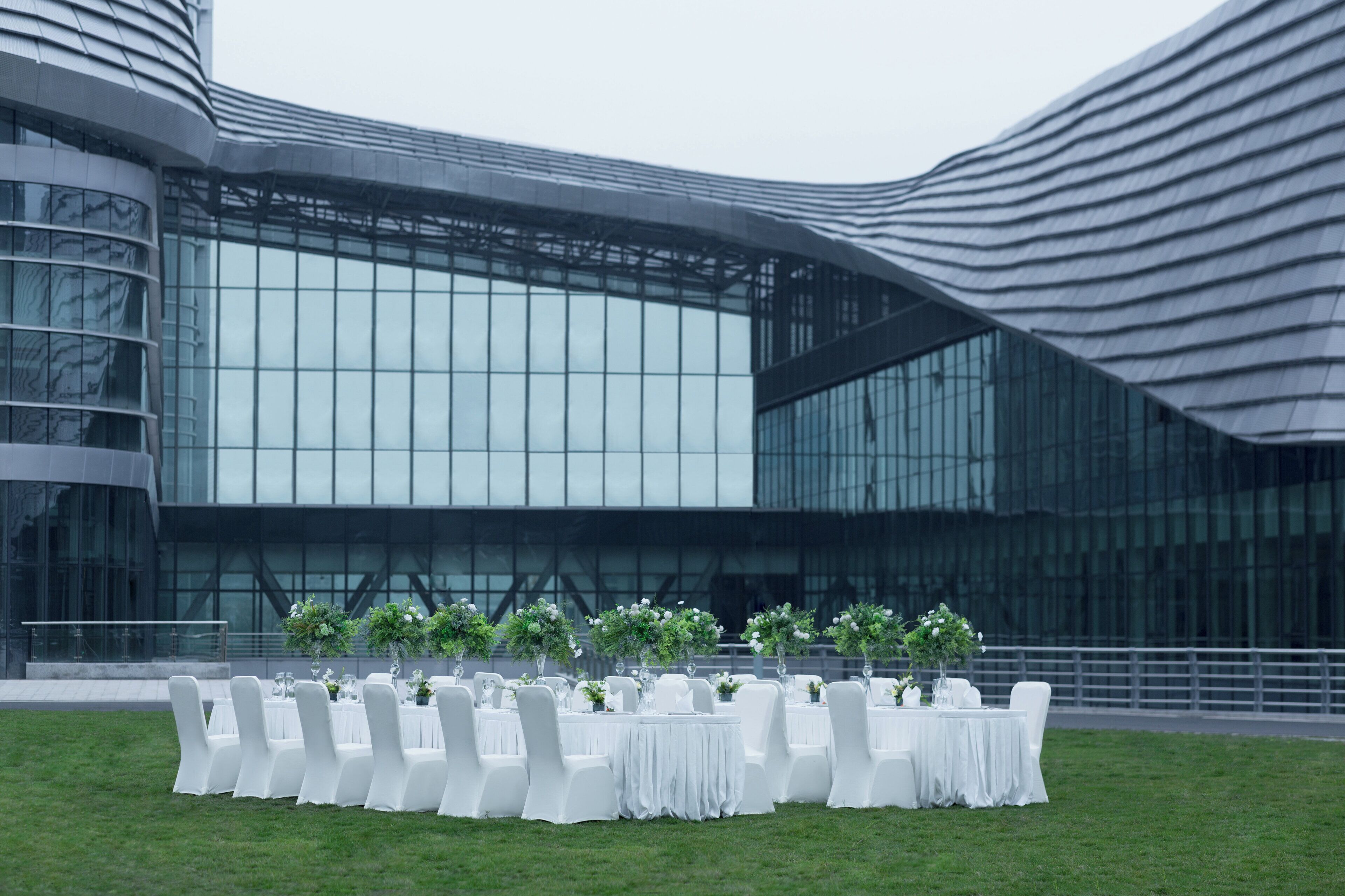 Un edificio moderno con fachada de vidrio curva y un área verde exterior con mesas redondas dispuestas para un evento.