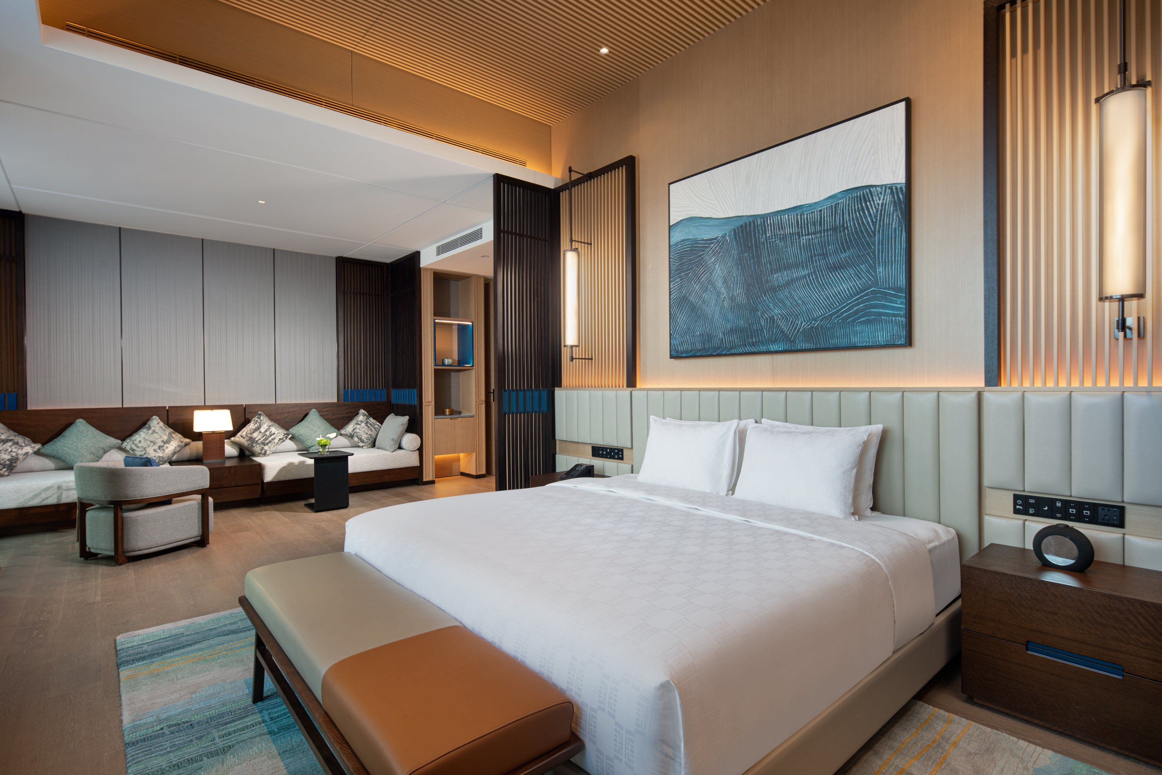 A modern hotel room with a large bed, a seating area with a sofa and armchairs, a bedside table with a lamp, and a large abstract painting on the wall.