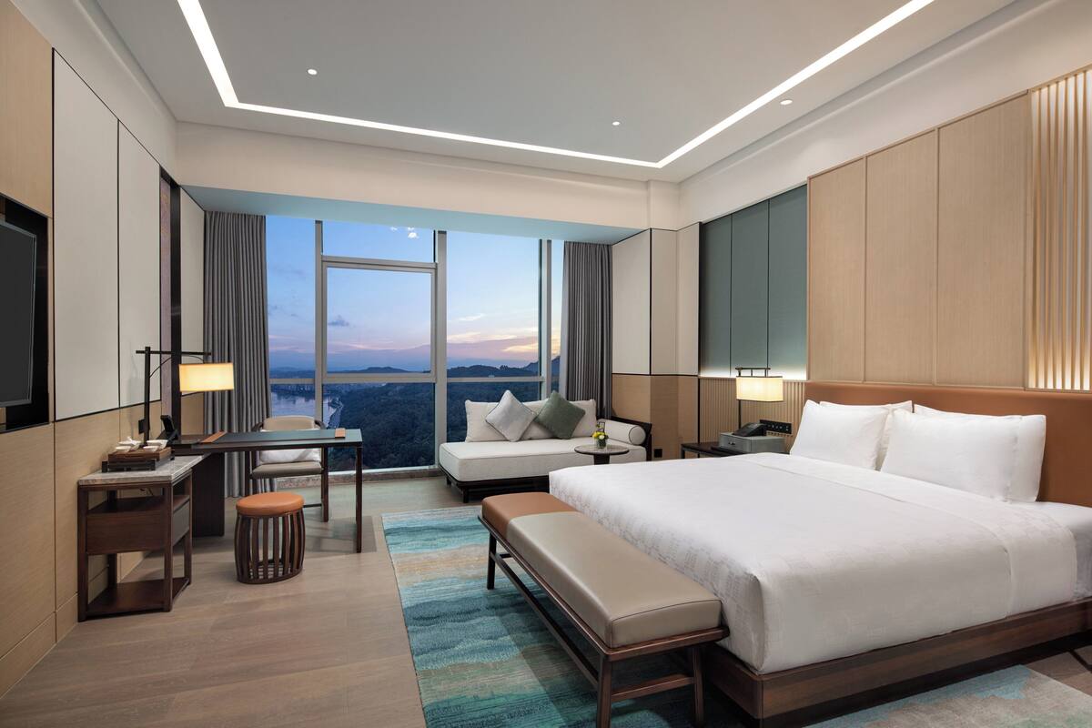 A modern hotel room with a large bed, a desk, a sofa, and a view of the cityscape.